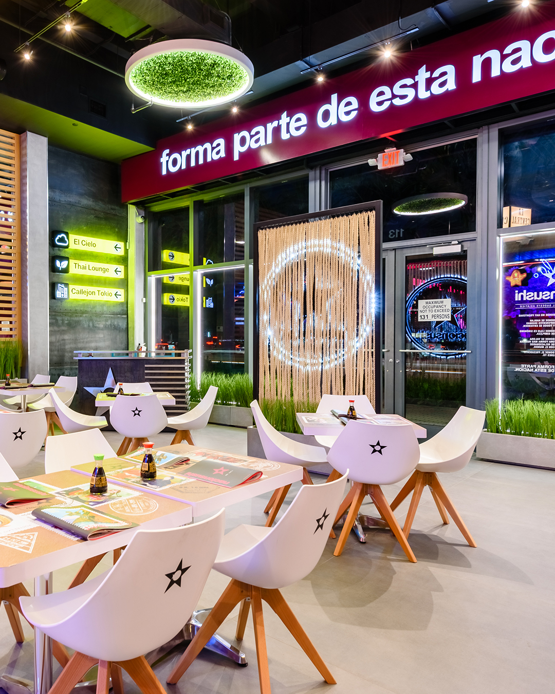 Área de mesas en Nacionsushi con sillas blancas, mesas de madera y señalización luminosa en el interior del restaurante.