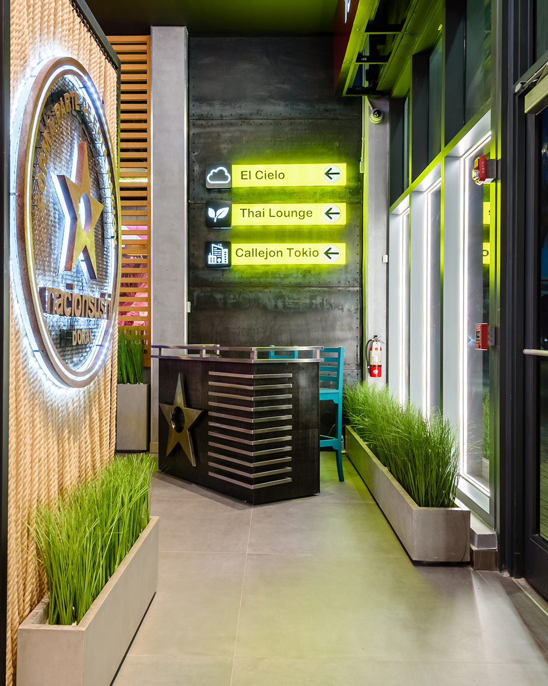 Área interior de Nacionsushi con mostrador de recepción, señalización luminosa hacia distintas zonas y jardineras decorativas.