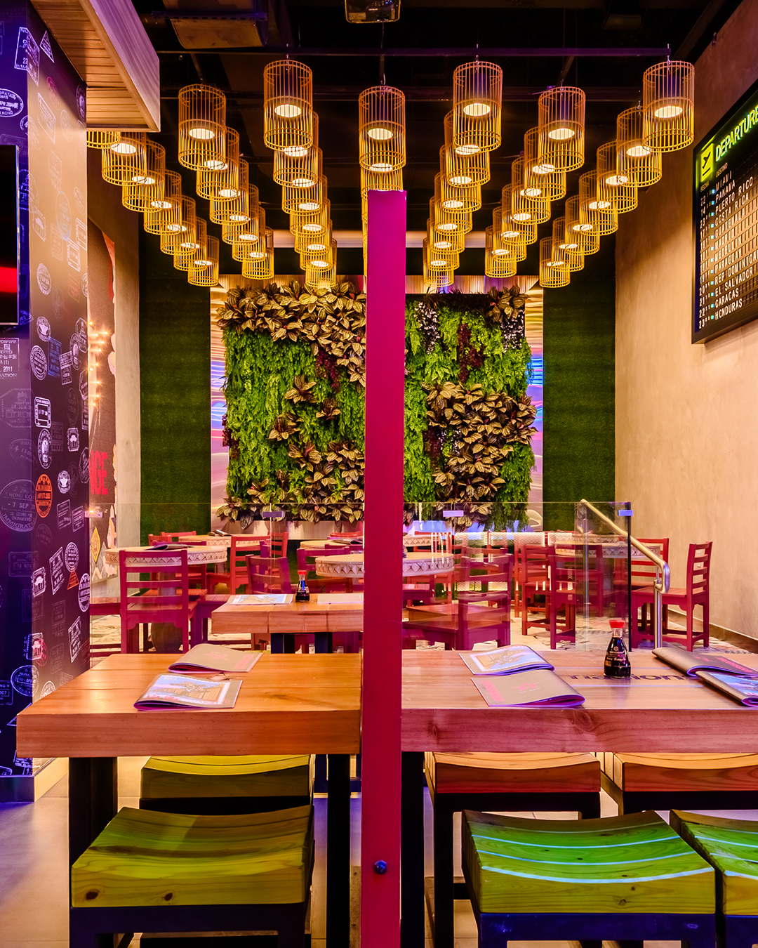 Área de mesas en Nacionsushi con mesas de madera, sillas coloridas, lámparas colgantes y muro vegetal decorativo.