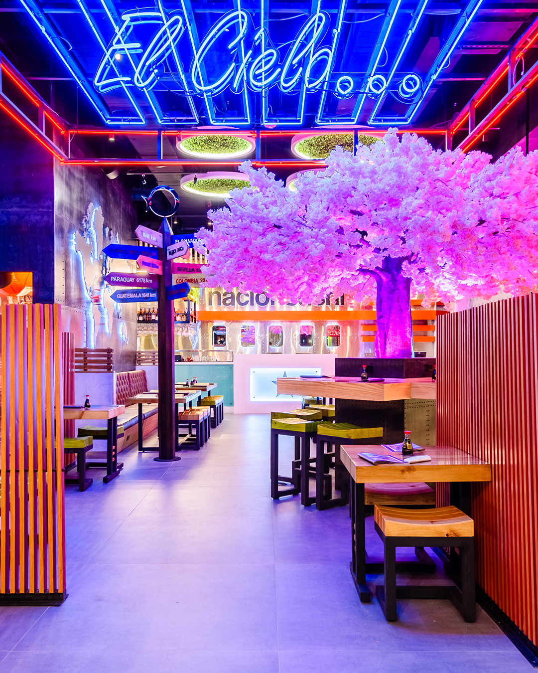 Vista interior de Nacionsushi con mesas de madera, árbol decorativo iluminado en tonos rosados y letrero de neón en el techo.